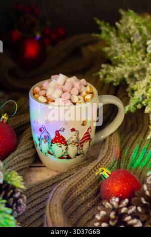 Ein warmer Becher mit Zwergen ist gefüllt mit farbenfrohen Marshmallows, die zwischen Tannenzapfen und Ornamenten ruhen und einen gemütlichen Urlaubsgeist im Innenbereich wecken. Stockfoto