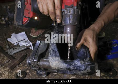 Fokussierter Mechaniker mit schmutzigen Händen, der Akku-Bohrmaschine für Reparaturarbeiten hält. Der Arbeiter bohrt vorsichtig ein Loch in das schwarze Kunststoffteil in der unordentlichen Werkstatt Stockfoto