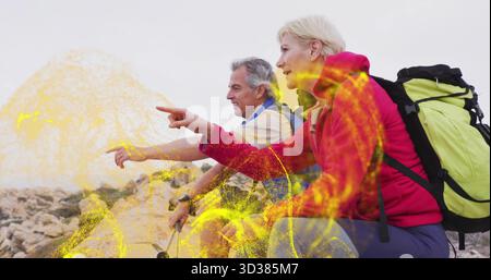 Zeigendes reifes Paar, das Jacken trägt, die auf dem Kamm nach vorne geneigt sind, mit Rucksäcken und Trekkingstöcken Stockfoto