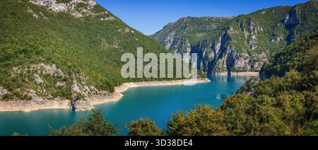 Panoramablick auf den Piva-See in Montenegro, umgeben von steilen grünen Bergen und felsigen Klippen unter klarem blauen Himmel, der sich in ruhigem türkisfarbenem Wasser spiegelt Stockfoto
