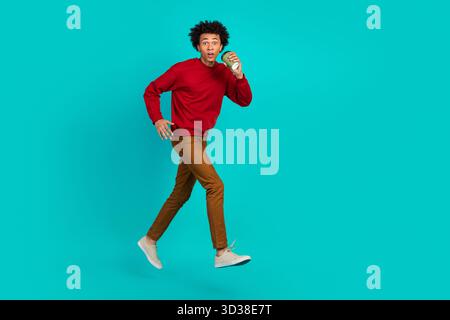 Junger Mann springt in leuchtend blauem Studio in rotem Pullover, braunen Hosen und weißen Sneakers, die eine Tasse in der Luft halten, stilvoll und lässig Stockfoto