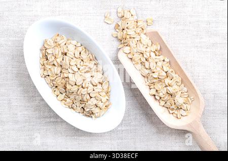 Haferflocken, dickgerollte, altmodische Haferflocken, in weißer Schüssel und Körnerlöffel, auf Leinen. Geschälte, gedämpfte Hafergrieß, zu Flocken gerollt und geröstet. Stockfoto