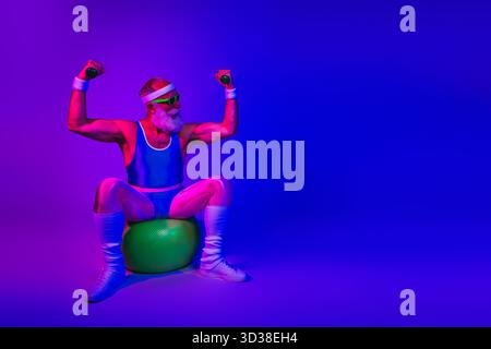 Ein bärtiger Seniorenmann im Retro-Fitness-Outfit trifft eine kräftige Pose auf einem Trainingsball unter neonvioletten und blauen Lichtern, die Kraft und Freude zeigen Stockfoto