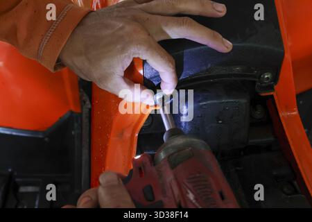 Fokussierte Mechaniker reparieren Fahrzeug mit Elektrowerkzeug für die Wartung. Mann bei der Arbeit, Befestigungsschraube an einer orangefarbenen Maschine mit einem Schlagschrauber in der Garage, s Stockfoto