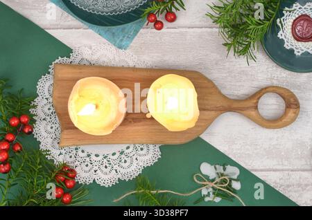Zwei brennende Kerzen auf Holzbrett, Weihnachtsdekoration, Häkelservietten, Briefmarken, Wachs, Teller, Nadelzweige und rote Beeren. Stockfoto