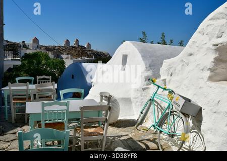 Amorgos, Griechenland - 11. September 2025: Farbenfrohe Sitzgelegenheiten und Fahrrad auf einer mit Steinen gepflasterten Terrasse mit Blick auf die alten Windmühlen auf dem Hügel Stockfoto