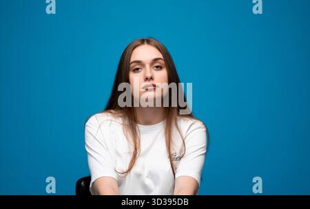 Neutrale Emotion einer jungen Frau mit geradem Gesicht in weißem T-Shirt auf blauem Hintergrund Stockfoto