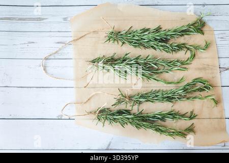 Vier Rosmarinbündel, zum Trocknen vorbereitet, gestylt auf Pergamentpapier mit hölzernem Hintergrund. Perfekt für saisonale Rezepte oder Kräuterhandwerk Stockfoto