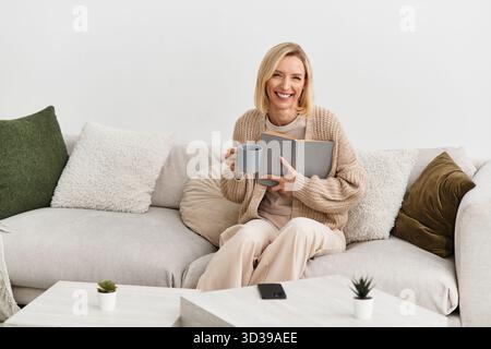 In einem modernen Apartment entspannt sich eine blonde Frau mit einem Buch und einem warmen Getränk und genießt ihren gemütlichen Raum. Stockfoto
