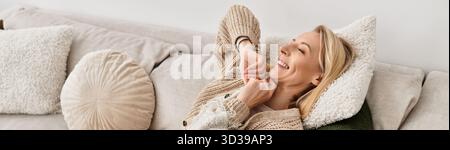 Die blonde Frau entspannt sich auf einer weichen Couch und genießt Wärme und Komfort in ihrem stilvollen Zuhause, Banner Stockfoto