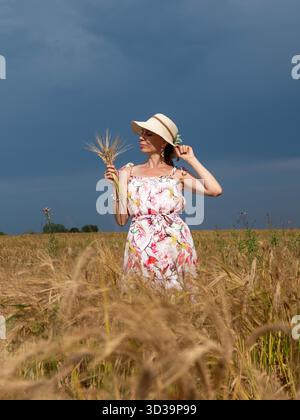 Eine vertikale Aufnahme einer schlanken Frau in den 40er oder 50er Jahren, die ein Sommerkleid aus Leinen, einen Hut und einen Haufen Getreide trägt, steht tagsüber mitten auf einem Feld. Ländliches Gelände mit landwirtschaftlicher Nutzung || Modell veröffentlicht Stockfoto