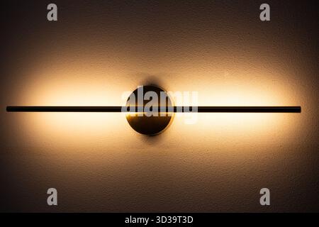 Diese moderne, minimalistische Wandleuchte mit linearem Design verleiht einer strukturierten Wand ein warmes, weiches Leuchten und schafft eine gemütliche Atmosphäre. Stockfoto