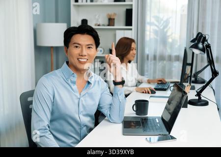 Ein intelligenter asiatischer Geschäftsmann, der mit seiner Kamera posiert und für Werbeflächen im modernen Büro nach oben zeigt. Verschwommener Hintergrund Frau, die arbeitet Stockfoto