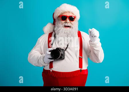 Weihnachtsmann mit Kamera in auffälliger Studioszene mit roten Hosenträgern und Hut hält Weihnachtsmomente für lustige Lifestyle-Fotos fest Stockfoto