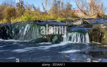 Kleiner Wasserfall mit sauberem Wasser und grünem Moos auf Steinen im Sonnenlicht Stockfoto