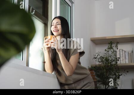 Eine Frau lächelt, während sie eine Kaffeetasse hält, aus dem Fenster blickt und einen Moment der Freude und Entspannung zu Hause verkörpert Stockfoto