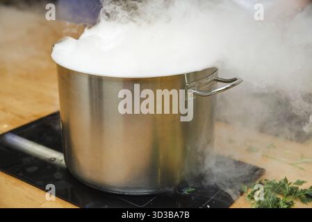 Lebensmittel, großer Topf aus Edelstahl auf dem Herd, der starken Dampf abgibt, bei intensivem Kochen überläuft, was auf einen aktiven Kochprozess in der Küche hinweist Stockfoto