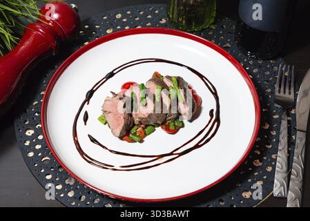 Essen, in Scheiben geschnittenes Rinderfilet, serviert mit Spargel und Kirschtomaten, künstlerisch mit balsamischer Reduktion auf einem weißen Gericht überzogen Stockfoto