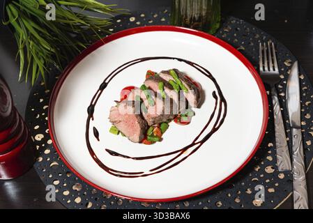 Speisen, überzogenes Gourmetgericht mit mittelseltenem Steak, gegrilltem Spargel, Kirschtomaten und Balsamico-Glasur auf einer weißen Keramikplatte Stockfoto