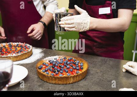 Essen, Konditor in Handschuhen streuen Puderzucker auf eine frische Fruchttorte, garniert mit Heidelbeeren und Erdbeeren während der Zubereitung Stockfoto