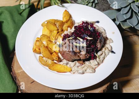 Speisen, saftiges gegrilltes Steak mit Preiselbeersauce, serviert mit gebratenen Kartoffeln und cremiger Pilzsauce auf einem weißen Teller Stockfoto