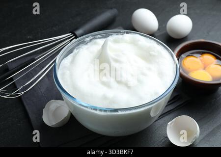 Eiweiß in Schüssel, Schneebesen und Zutaten auf schwarzem Tisch, Nahaufnahme Stockfoto