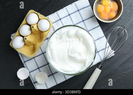 Eiweiß in Schüssel, Schneebesen und Zutaten auf schwarzem Tisch, flach gelegt Stockfoto