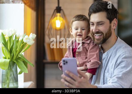 Bärtiger Vater, der mit der Tochter und dem Mobiltelefon steht und Videoanrufe oder Selfie in einem modernen Wohnraum macht Stockfoto