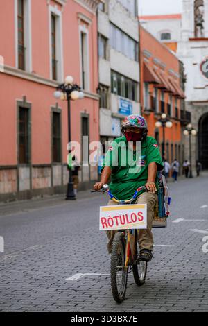 Mann mittleren Alters, der auf einem Arbeitsrad mit einem „RÓTULOS“-Schild und einer Schutzmaske auf einer kopfsteingepflasterten Straße in einer historischen Innenstadt fährt. Stockfoto