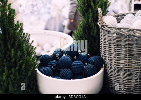 Blauer glitzernder Weihnachtsschmuck in einer weißen Schüssel zwischen künstlichen Kiefern und Korbkörben. Große Weihnachtsdekoration, Schneeballschmuck Stockfoto