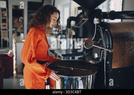 Junge Arbeiterin in orangefarbenem Anzug in Werkstatt mit Kaffeeröstermaschine während des Kaffeerösterprozesses Stockfoto
