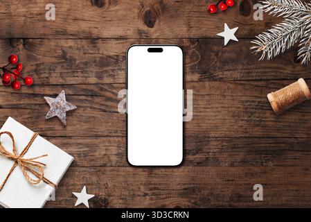 Smartphone mit leerem weißen Bildschirm auf rustikalem Holzhintergrund, umgeben von Weihnachtsdekorationen, Kiefernzweigen und festlichen Ornamenten Stockfoto