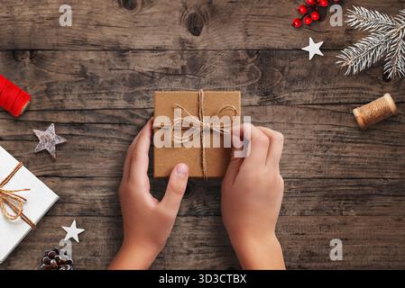 Hände halten umweltfreundliche Weihnachtsgeschenk-Box in braunem Papier und Garn auf rustikalen Holztisch mit festlicher Dekoration Stockfoto