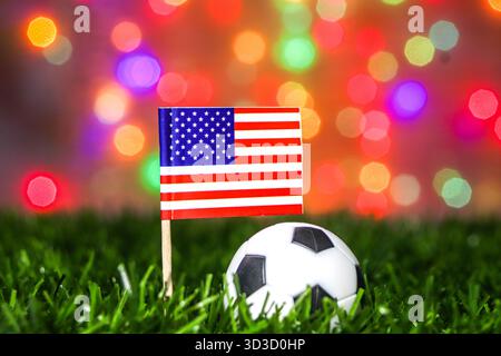 Nahaufnahme der amerikanischen Flagge und des Fußballballs auf dem Rasen mit einem hellen, abstrakten Hintergrund mit verschwommenen Regenbogenlichtern. Symbolisiert den globalen Sport. Stockfoto