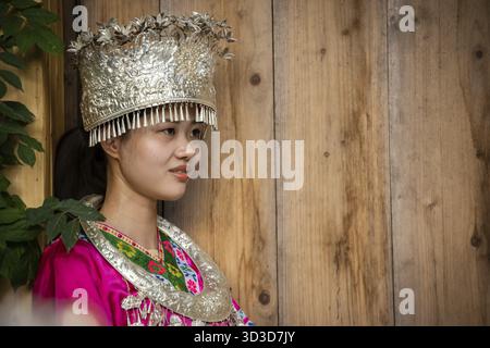 Feng Huang, China - August 2019 : schöne Chinesin in traditioneller Volkstracht gekleidet und posiert für ein Bild vor hölzernem Hintergrund von an Stockfoto