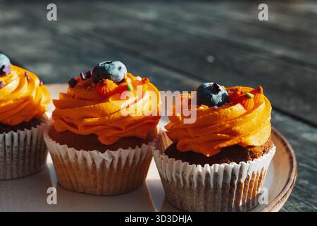 Köstliche Cupcakes mit Orangenfrost und Heidelbeerbelag auf einem Teller auf einem rustikalen dunklen Holztisch. Diese leckeren Süßigkeiten und Leckereien sind perfekt für Stockfoto