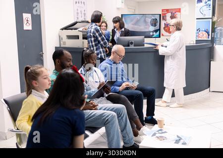 Hausärztin mit Klemmbrett sitzt zwischen älteren Mann und jungen schwarzen Mann in der klinischen Lobby. Diverse Patienten warten in der Nähe auf ärztliche Konsultationen im modernen Wartezimmer des Krankenhauses. Stockfoto