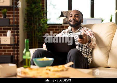 afroamerikanischer Freelancer arbeitet ferngesteuert vom Sofa aus mit einem digitalen Gerät auf dem Schoß und stellt die Lautstärke des Fernsehers über die Fernbedienung ein. Junger Mann Multitasking zwischen Laptop und Fernsehen zu Hause. Stockfoto
