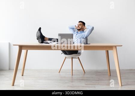 Ein lächelnder arabischer Mann, der nach der Arbeit an seinem Schreibtisch Entspannung genießt Stockfoto
