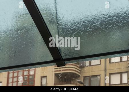 Starker Regen stürzt über ein Glasdach, Tröpfchen strömen den Schutzraum hinunter mit verschwommenen städtischen Gebäuden im Hintergrund. Stockfoto