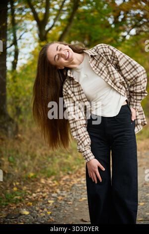 Eine junge Frau mit langen Haaren lächelt hell, während sie ein kariertes Hemd und eine schwarze Hose trägt. Sie steht auf einem Pfad, umgeben von bunten Herbstbäumen im Park. Stockfoto