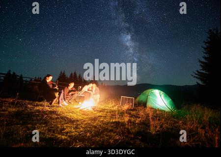 Eine Gruppe von Wanderfreunden ruht am Lagerfeuer neben einem beleuchteten Zelt und einem Solarpanel unter sternenüberzogenem Himmel. Die Milchstraße leuchtet hell über den Bergen und schafft eine magische Atmosphäre in der Wildnis der Berge. Stockfoto