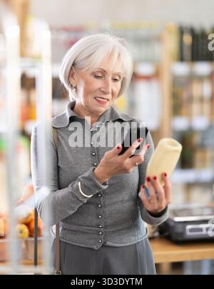 Ältere Käuferinnen scannen QR-Code auf Flaschenmayonnaise-Etikett in der Lebensmittelabteilung des Supermarktes Stockfoto