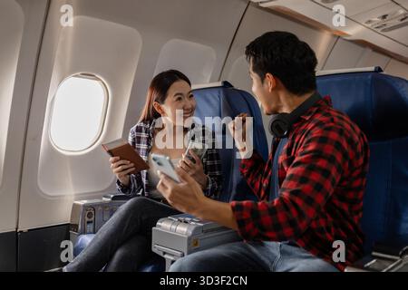 Das aufregende junge asiatische Paar sitzt in einem Flugzeug, hält einen Pass, Bargeld und Smartphone und feiert zusammen. Die Frau sieht glücklich aus und die Stockfoto