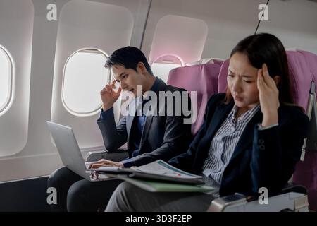 Zwei gestresste asiatische Geschäftsleute sitzen in einem Flugzeug und halten den Kopf frustriert. Der Mann schaut auf den Laptop-Bildschirm, während die Frau Stockfoto