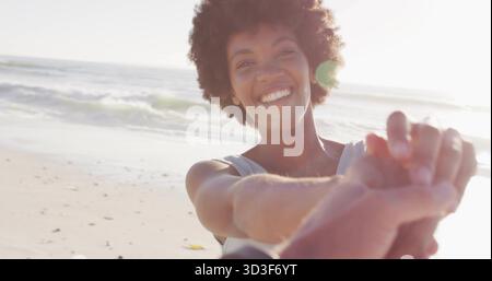 Lächelnde Schwarze Frau mit ärmellosem Oberteil, die Hand am Sandstrand ausstreckt, mit Meereswellen Stockfoto