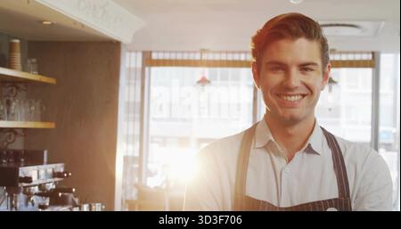 Ein lächelnder Barista bereitet Espresso an der Kaffeekasse mit Mahlwerk, Glasbechern und Hängeleuchten zu Stockfoto