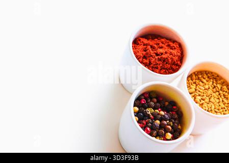 Drei weiße Schüsseln sind mit verschiedenen Gewürzen gefüllt, darunter rote Pfefferflocken, Pfeffer und gelbe Samen vor einem sauberen weißen Hintergrund. Stockfoto