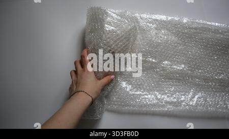 Weibliche Hand rollt ein großes Blatt Luftpolsterfolie auf weißem Hintergrund auf. Konzept der Verpackung zerbrechlicher Gegenstände, Schutz für den Versand und Lieferung von g Stockfoto