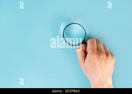Die Hände des Mannes halten eine Lupe auf dem blauen Tischhintergrund. Der Begriff der sorgfältigen Betrachtung und Suche. Nahaufnahme, POV-Aufnahme. Stockfoto